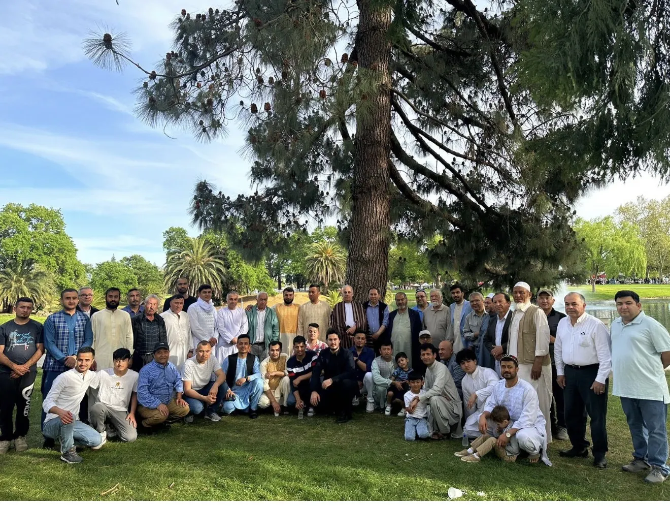 Members of the Afghan Turkic Community gathered together beneath a tall pine tree in a California park