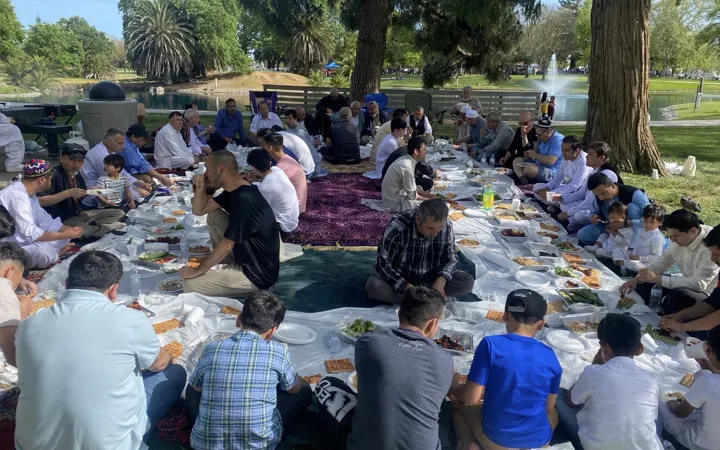 Community members sitting on carpets in a park, sharing a large picnic meal